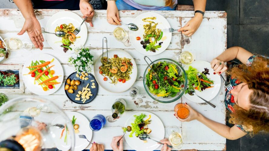 A white dining table shown from overhead with multiple women sitting around it and eating from plentiful bowls of salads and other Mediterranean diet foods.