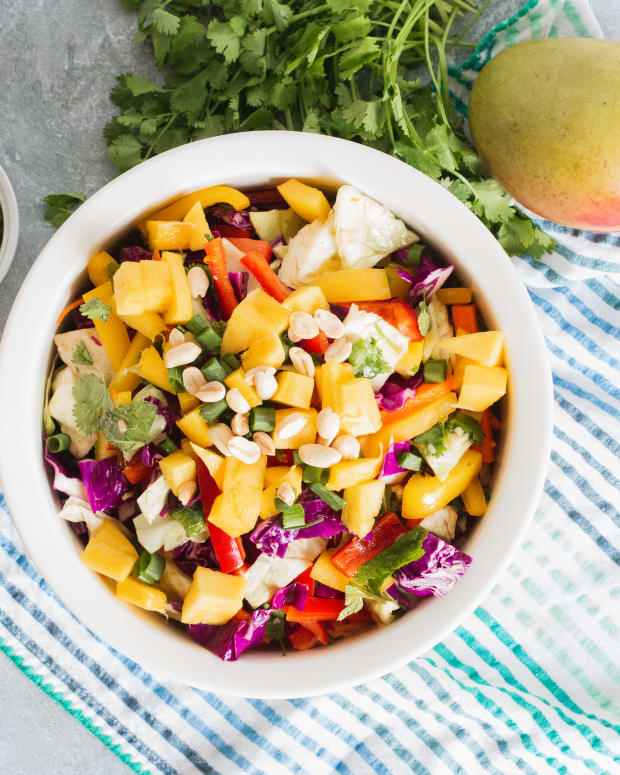 spicy mango asian salad in a white bowl surrounded by peanuts green onions, cilantro and a whole mango.