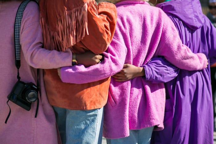 Four women holding each other wearing colorful pink, purple, and orange coats.