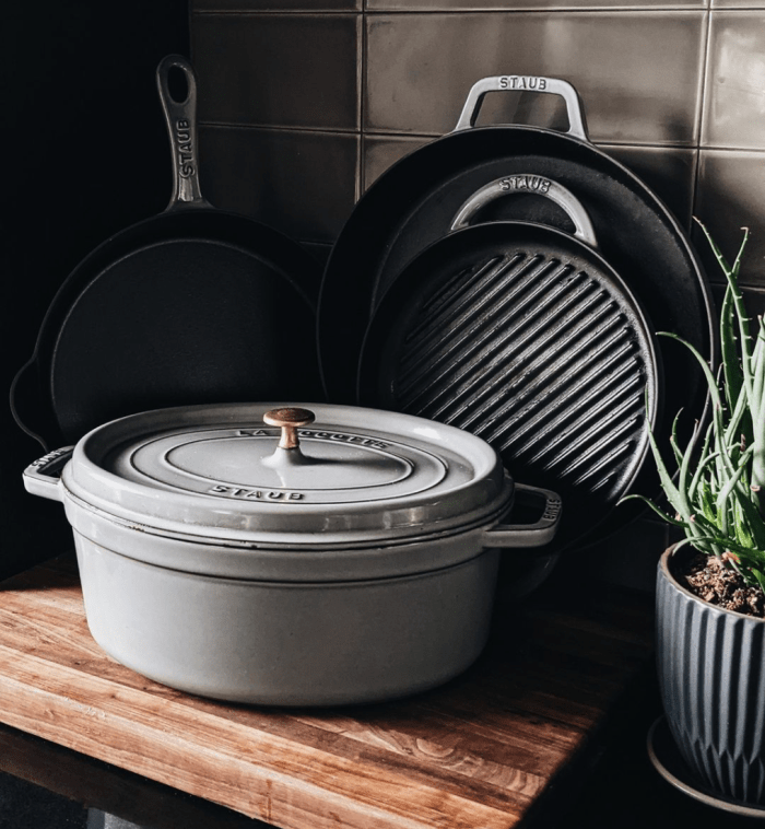 Staub cast iron oval cocotte in graphite grey in front of cast iron grill and saute pans.