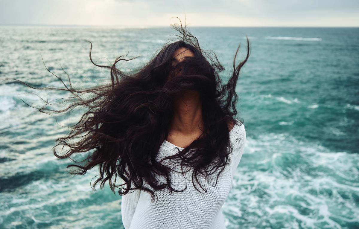 Woman with long hair stands in the wind in front of the ocean.