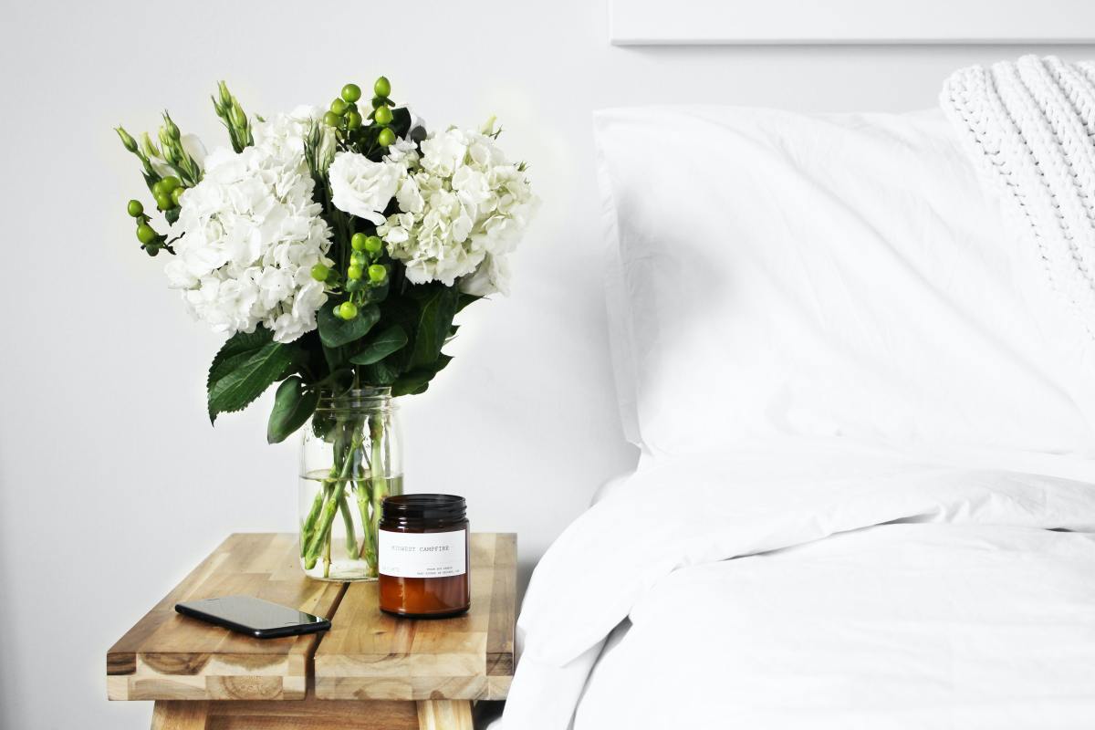 White bouquet, candle, and phone next to bed with white sheets.