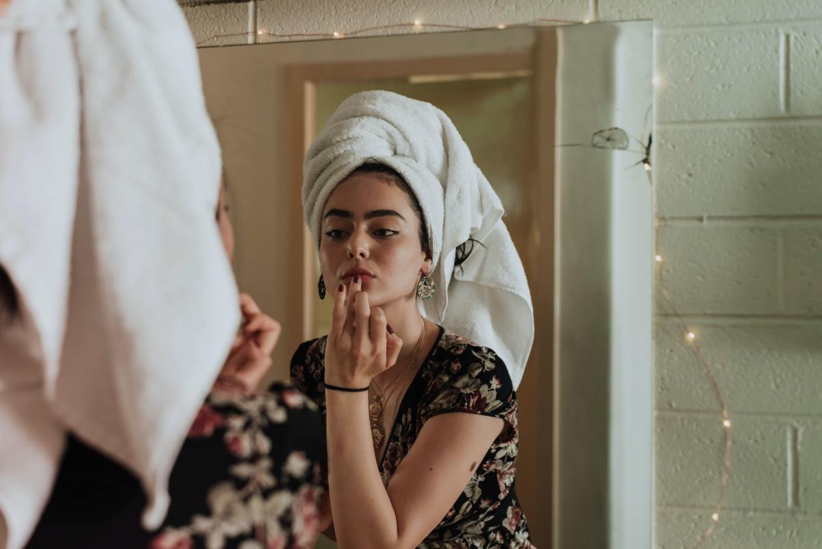 Woman applies makeup and skincare in mirror with towel on her head.