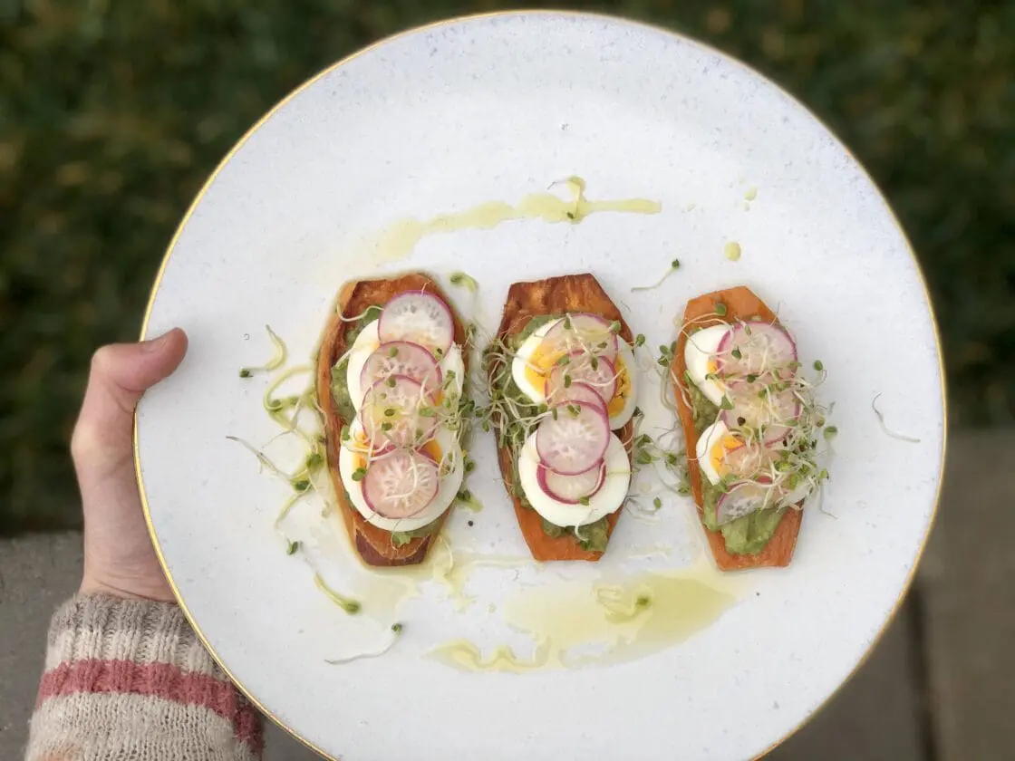 Hand holding plate of sweet potato toast with hard boiled egg, sprouts and avocado.