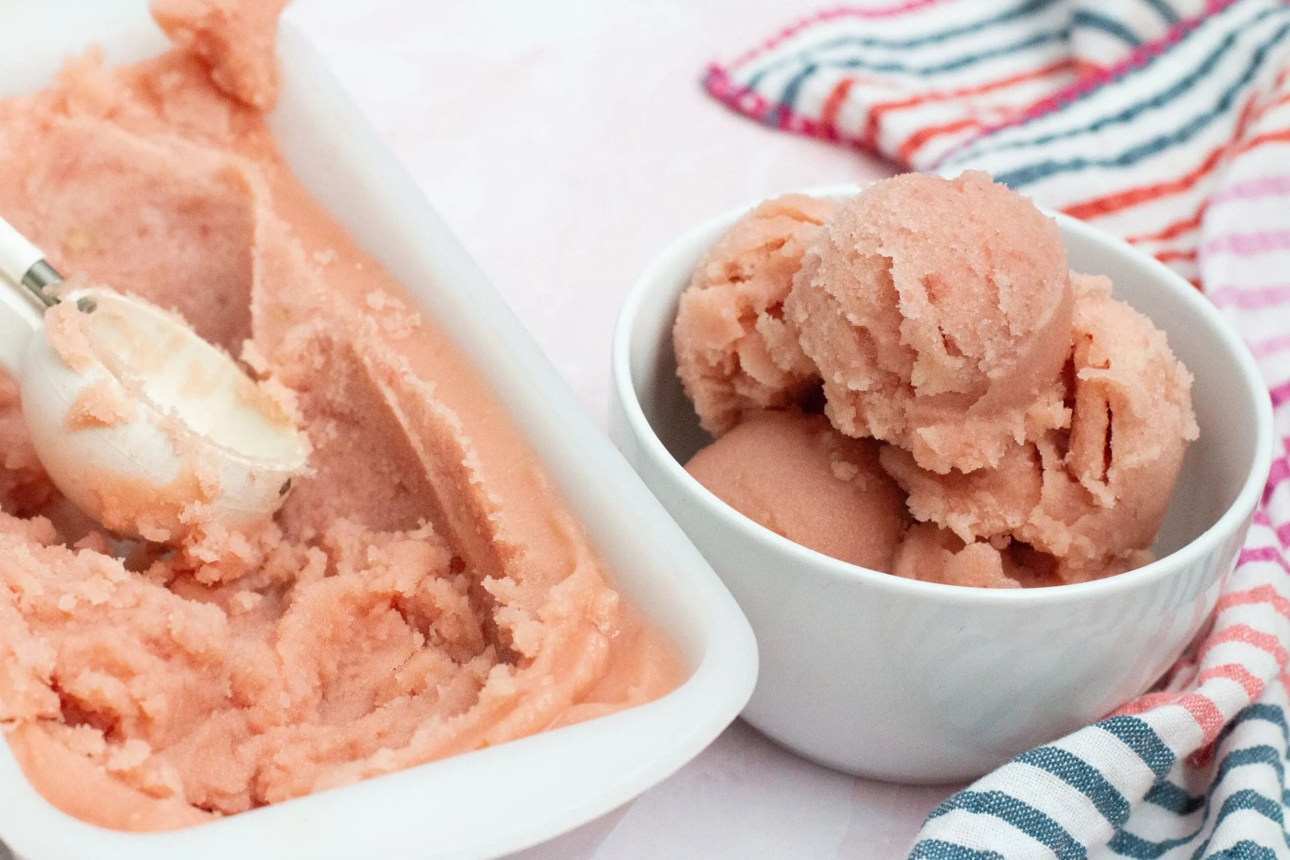 Image: Scoops of pink watermelon sorbet in a white bowl using white ice cream scoop and out of a white enamel bread pan with colorful striped towel off to right side.