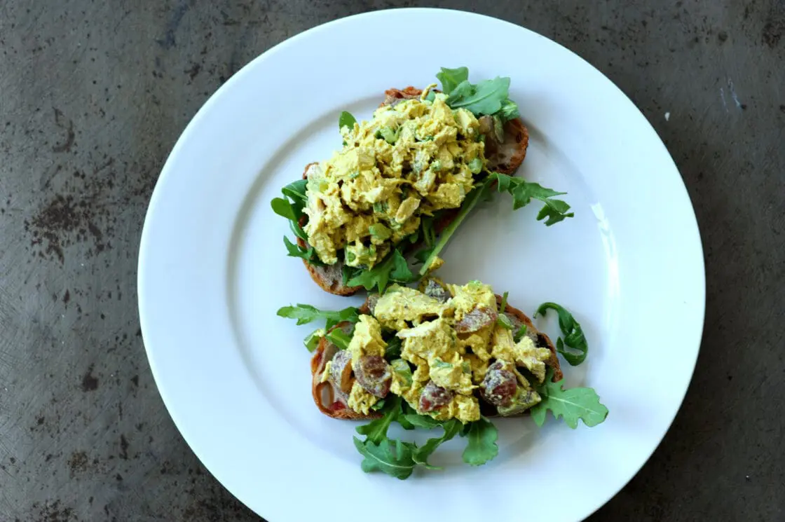 Image of an overhead view of a curry chicken salad recipe served with arugula over sourdough bread on a white plate and a concrete countertop.