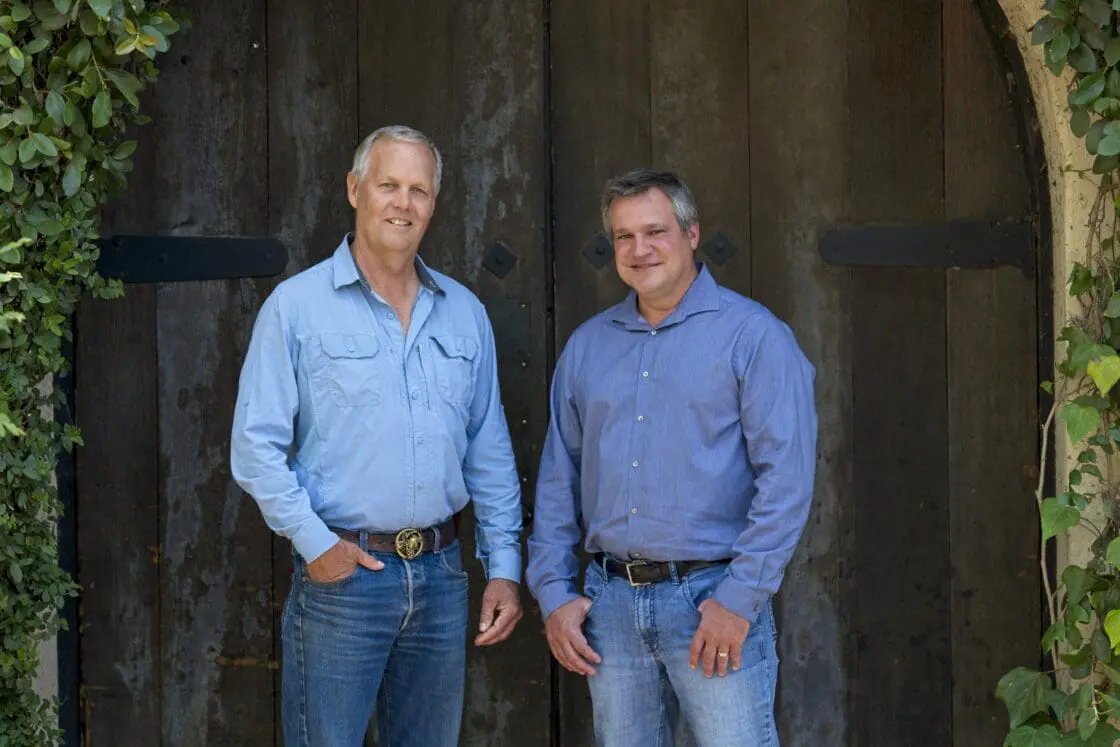 Image of Stag's Leap Wine Cellar's Vineyard Director, Kirk Grace, and Winemaker Marcus Notaro both wearing jeans and blue button-down shirts, stand in font of an old, worn door with vines trailing over.