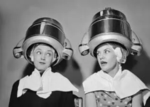 Two women wearing large vintage hair dryer bonnets, looking upward in surprise.