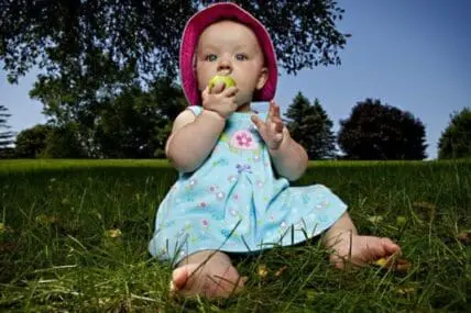 Baby sitting on grass in a blue dress and pink hat, eating an apple