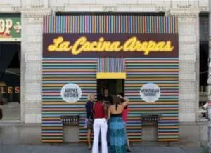 Three people standing in front of La Cocina Arepas storefront with colorful striped facade.