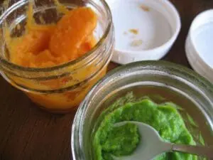 Two glass jars containing homemade baby food purees in orange and green colors with spoons