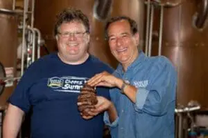 Two men posing together in a brewery with large copper tanks in the background