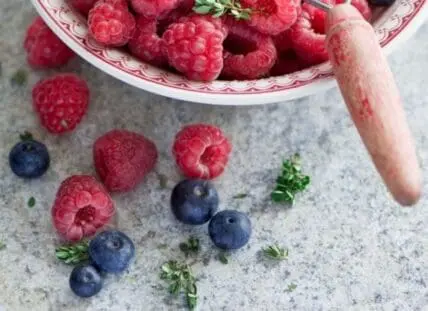 Fresh raspberries and blueberries in a decorative bowl with herbs and a pestle on stone surface