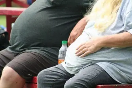 Two overweight people sitting on a red bench with a water bottle between them