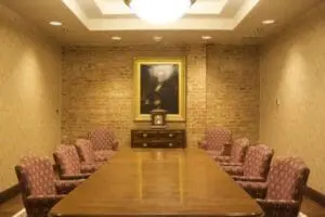 Formal boardroom with long wooden table, red patterned chairs, and portrait painting on brick wall