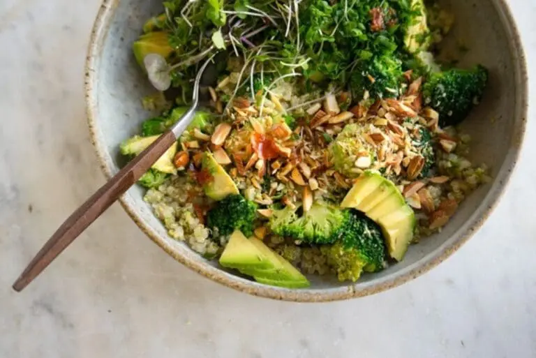 Image of overhead view of a broccoli Buddha bowl served in a speckled bowl, with a wood handled spoon on a marble surface with a sprinkle of broccoli sprouts nutrition
