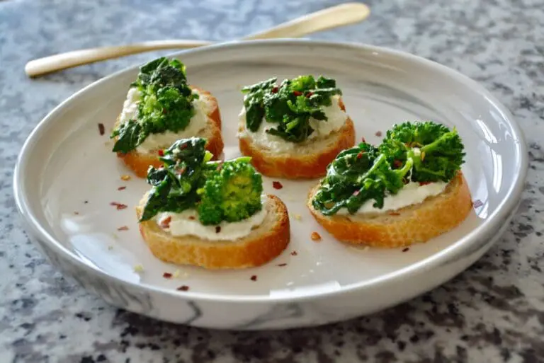 Image of 2 broccoli rabe crostini on a white plate on a marble surface with gold utensil in the background showing broccoli sprouts nutrition