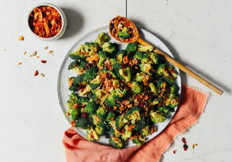 Image of broccoli salad topped with chilis on a plate, styled with a peach kitchen towel, a wooden spoon, and a small white bowl of toppings for this sulforaphane broccoli sprouts packed powerhouse salad.
