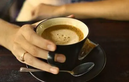 Person holding a black cup of coffee with saucer and spoon