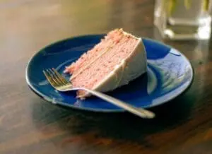 A slice of pink layer cake with white frosting on a blue plate with a fork.