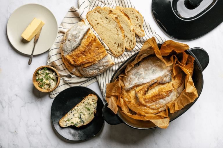 Image of a 100% ceramic cookware, safe Dutch oven filled with crusty bread cooked to golden perfection in unbleached parchment paper. The lid of the black Xtrema Dutch oven sits off to the side on a white marble work surface with a white kitchen towel with gray stripes, a small white plate with a stick of butter and butter knife, a small black plate with a slice of fresh bread smeared with herb butter.