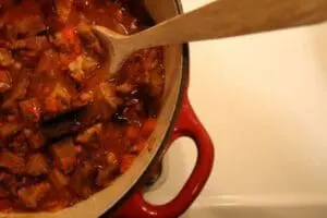 A red Dutch oven filled with pumpkin chili being stirred with a wooden spoon.