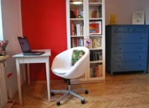 Modern home office with red accent wall, white desk, curved chair, blue dresser, and built-in