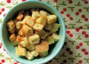 Homemade croutons in a turquoise bowl on a cherry-patterned cloth