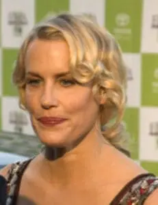 Woman with blonde wavy hair at a media event with green and white backdrop