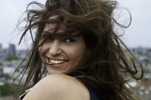 Woman with long, windswept brown hair smiling outdoors with city skyline behind her