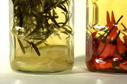 Two glass jars with herb-infused vinegars, one pale yellow and one deep red, with fresh herbs