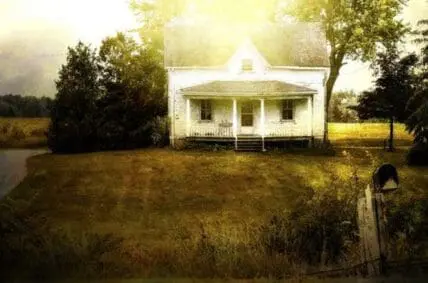 Abandoned white farmhouse with front porch surrounded by overgrown fields and trees