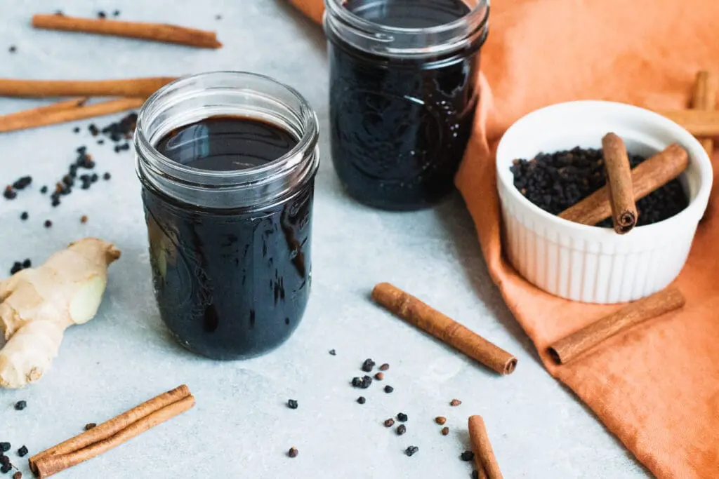 image of homemade elderberry syrup for cold and flus in mason jars with a white ramekin of elderberries and cinnamon sticks with an orange kitchen towel.