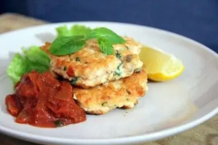 Two crispy salmon patties stacked on a white plate with lemon wedge and red sauce