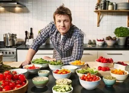 Man in plaid shirt surrounded by fresh vegetables and ingredients in a kitchen.