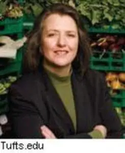 Woman in black blazer and olive shirt smiling outdoors with greenery background
