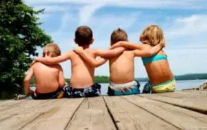 Four children sitting together on a wooden dock by a lake with their arms around each other
