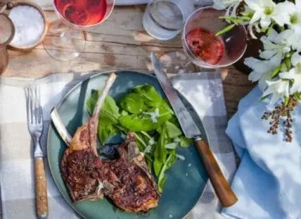 Plated lamb chops with fresh greens and mint sauce beside wine glasses and flowers