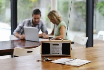 Portable speaker and notepad on desk with two people working on laptop in background