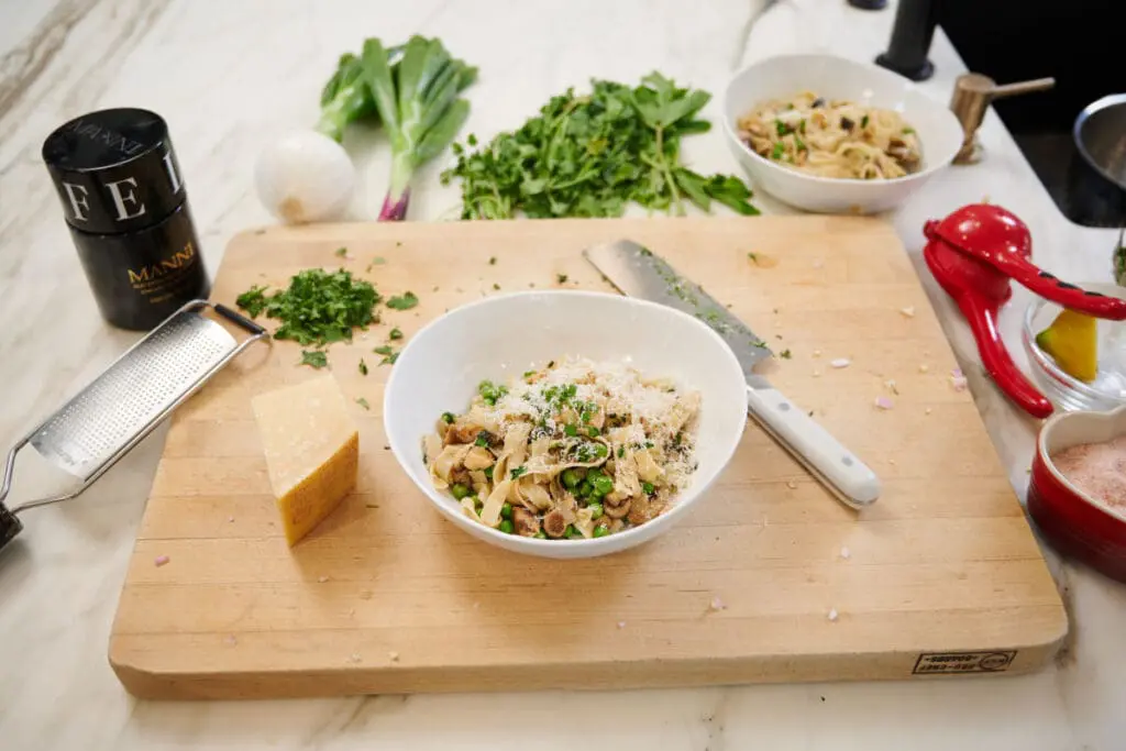 Image of a white bowl of lions mane pasta recipe with fresh peas, herbs, and parmesan served on a wood cutting board next to white-handled chef's knife and a block of parmesan cheese.