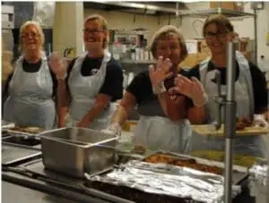 Four people in kitchen aprons and gloves prepare food in a school kitchen setting.