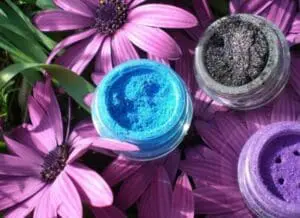 Three loose mineral makeup powders in blue, black, and purple displayed on purple flowers