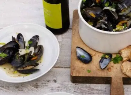 Cooked mussels in white wine sauce with bread and wine bottle on wooden surface