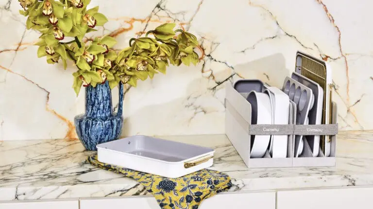 Image of a Caraway clean, non-toxic bakeware stored on modern kitchen counter with marble and marble backsplash. One bake pan sits out on a dish towel, and the rest are stored in Caraway Home's storage containers.