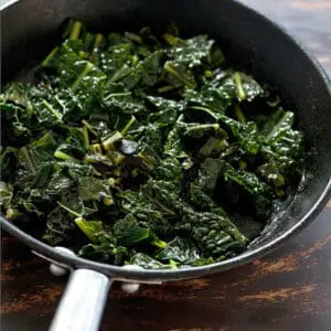Braised dark leafy greens cooking in a black skillet with glistening moisture