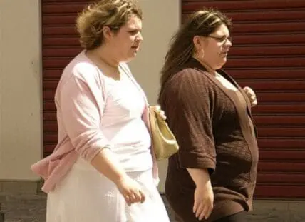 Two women standing together against a wall with red shutters in the background.