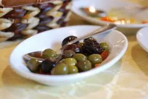 A white bowl of mixed green and black olives with a serving spoon on a table.