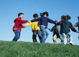 Children holding hands in a circle on a grass hill against a blue sky