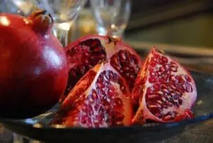 Whole and halved pomegranates displaying seeds on a dark plate with glasses in background