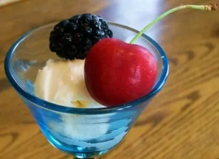 A glass dessert cup filled with whipped cream, topped with a red cherry and dark berry